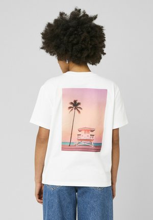 Personne aux cheveux bouclés portant un t-shirt blanc avec un imprimé coucher de soleil d'une tour de sauveteur et un palmier, associé à un jean bleu.