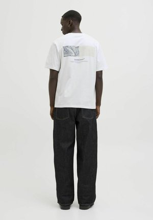 T-shirt blanc à manches courtes avec un imprimé graphique minimaliste au dos, associé à un jeans sombre ample et des chaussures noires.