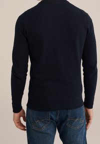 Man i en mörk marinblå strukturerad tröja och blå jeans, vänd bort från en enfärgad beige bakgrund.