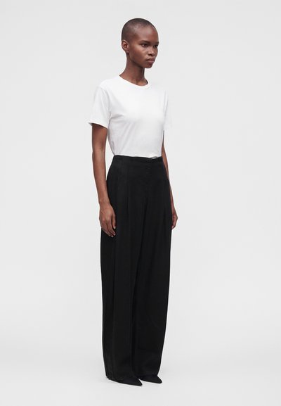 Personne aux cheveux courts portant un haut blanc à manches courtes et un pantalon large noir taille haute, debout sur un fond clair uni.