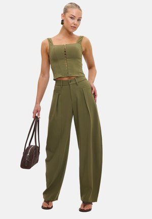Femme portant un débardeur court vert olive et un pantalon large taille haute, tenant un sac à main marron avec des sandales sur fond blanc.