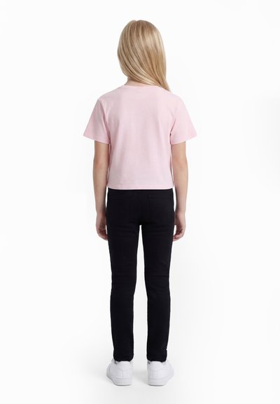 Dijete stoji okrenuto leđima, odjeveno u svijetloružičastu majicu kratkih rukava, crne hlače i bijele tenisice na bijeloj pozadini.