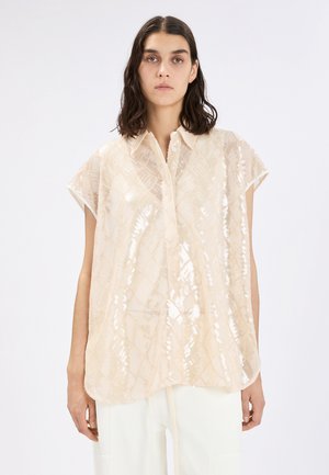 Donna con capelli scuri di media lunghezza che indossa una blusa beige a maniche corte e larghe con paillettes e pantaloni bianchi, su uno sfondo neutro.