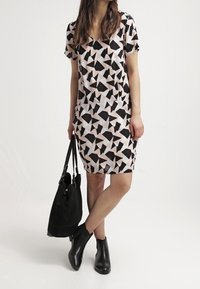 Geometrisk mönstrad klänning i mjuk rosa, svart och vit. Korta ärmar och V-ringning. Modellen bär en svart tote och har på sig ankelboots.