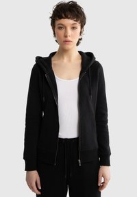 Młoda osoba z krótkimi kręconymi włosami, ubrana w czarną bluzę z kapturem na zamek, biały top na ramiączkach i czarne spodnie ze sznurkiem, stojąca na jasnym tle.