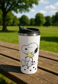 Tazza da viaggio bianca con coperchio nero che raffigura Snoopy che abbraccia Woodstock, posizionata su una panchina di legno in un parco verde sotto un cielo blu.