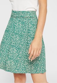 Zelená sukně s texturovaným vzorem bílých teček, vybavená elastickým pasem a rozšířeným střihem. Materiál vypadá lehce a prodyšně.