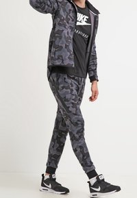 Camo-mönstrad träningsoverall i nyanser av grått, bestående av en jacka med dragkedja och ribbade muddar, kombinerad med matchande avsmalnande byxor och atletiska sneakers.