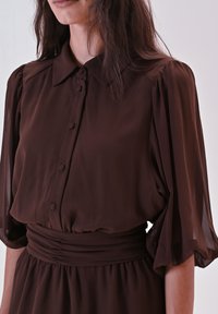 Donna che indossa un abito marrone scuro con maniche a sbuffo a tre quarti, bottoni rivestiti in stoffa e una ampia fascia arricciata in vita.
