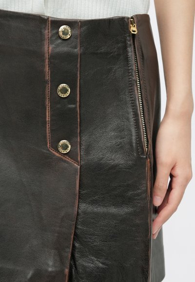 Jupe crayon en cuir noir avec une fermeture éclair et des accents de boutons dorés. Présente des coutures contrastées en brun et un design ajusté.