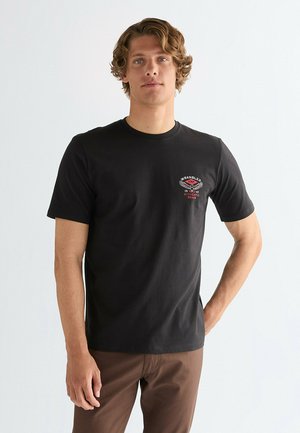 Zwart katoenen T-shirt met korte mouwen, ronde hals en een logo op de linkerborst. Model draagt lichtbruine broek en staat tegen een lichte achtergrond.