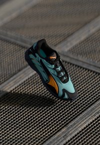 Tyrkysová síťovaná teniska s černými akcenty a oranžovými detaily, s elegantním designem a texturovanou podrážkou, umístěná nad mřížkovým povrchem.