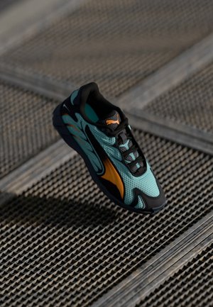 Turkusowe siateczkowe buty sportowe z czarnymi akcentami i pomarańczowymi detalami, o eleganckim designie i teksturowanej podeszwie, umieszczone nad powierzchnią w kratkę.