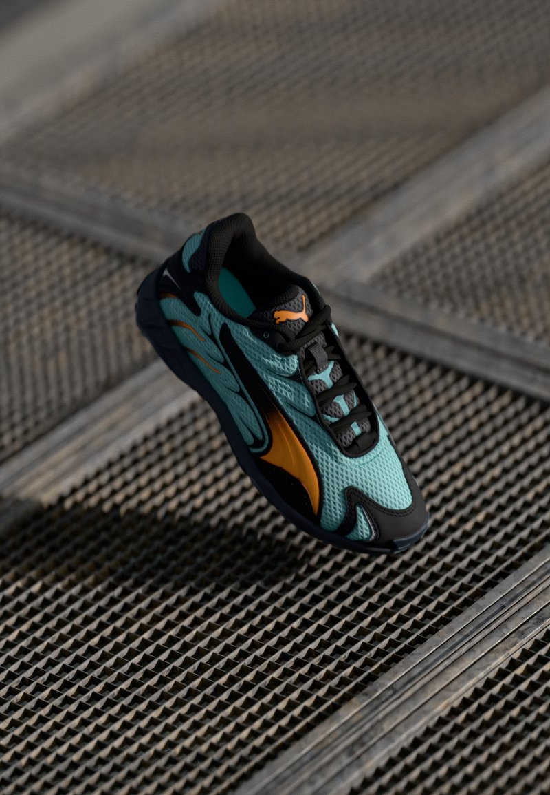 Tyrkysová síťovaná teniska s černými akcenty a oranžovými detaily, s elegantním designem a texturovanou podrážkou, umístěná nad mřížkovým povrchem.