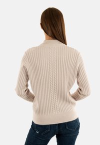 Maglione beige a coste con dettagli a trecce lungo la spina dorsale, dotato di scollatura rotonda e maniche attillate, abbinato a jeans scuri.