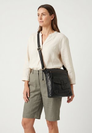Vrouw draagt een crèmewitte blouse en olijfgroene korte broek met een zwarte leren schoudertas, staand tegen een effen lichte achtergrond.