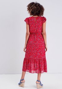 Robe maxi rouge à fleurs avec des manches volantées, taille cintrée et ourlet en étages. Le tissu est léger, avec un motif floral subtil.