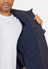 Laivaston takki, jossa vasemmalla sisäpuolella on vetoketjullinen tasku. Sileä, kestävä kangas ja irrotettava huppu. Sisäpuoli vuorattu vaaleammalla sinisellä.