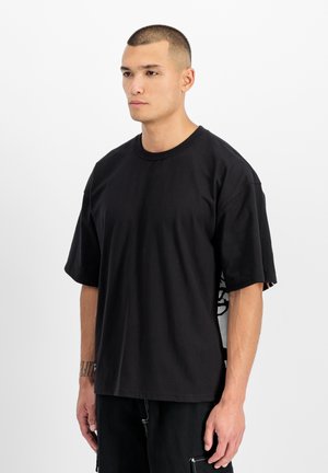 Young man with short hair wearing a loose black t-shirt and black pants, standing against a plain white background, looking slightly to the side.