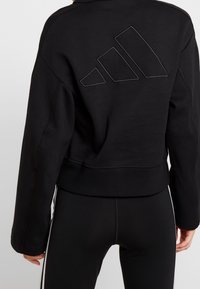 Sudadera negra con un diseño corto, que presenta un destacado logo bordado de Adidas en la parte trasera. Textura suave y puños acanalados.
