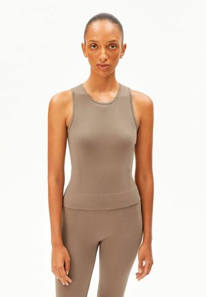 Frau steht frontal und trägt ein enganliegendes, ärmelloses Top in Taupe sowie passende Leggings vor einem schlichten weißen Hintergrund.