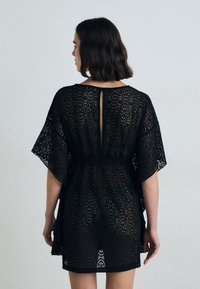 Abito in pizzo nero con un motivo geometrico, maniche corte e vita arricciata. Sul retro c'è un'apertura piccola all'altezza del collo.