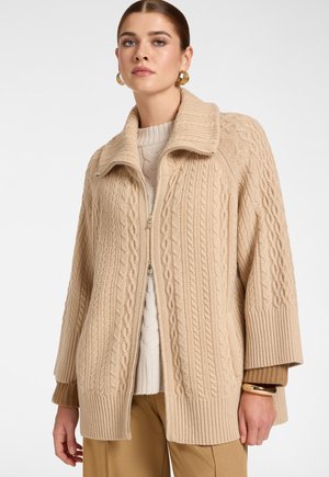 Femme portant un cardigan beige en maille torsadée zippé avec un large col, superposé à un pull crème et un pantalon marron, avec des boucles d'oreilles créoles dorées.