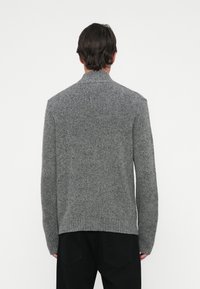 Mann, der von hinten schaut, trägt einen grauen Strickpullover und schwarze Hosen vor einem schlichten weißen Hintergrund.