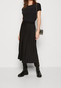 Falda negra plisada con cinturilla elástica, combinada con una blusa negra de manga corta que presenta acentos de strass. Botas negras hasta la rodilla y un bolso.