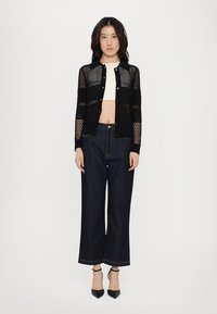 Cardigan corto in maglia trasparente nera con chiusura a bottoni, indossato sopra una maglietta bianca. Abbinato a pantaloni di jeans a gamba larga scuri e tacchi neri.