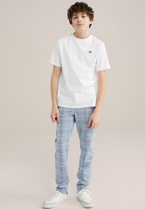 Weißes T-Shirt mit kurzen Ärmeln und einem kleinen schwarzen Logo, kombiniert mit hellblauen karrierten Hosen und weißen Sneakers mit blauen Akzenten.