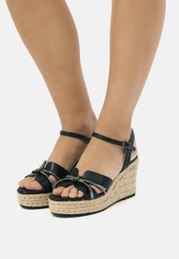 Sandali con zeppa nera dal design espadrille, caratterizzati da una suola in corda. Presentano cinturini intrecciati e un accento decorativo in argento sul davanti.