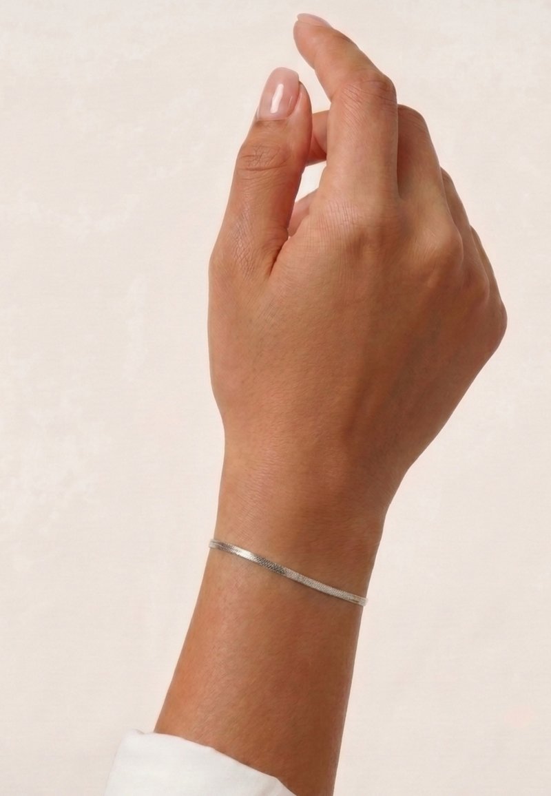 Main levée avec des ongles naturels portant un fin bracelet en argent sur un fond clair et uni. Manche blanche visible au poignet.