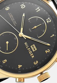 Cadran noir avec des accents en or, présente des index horaires, des sous-cadrans pour le jour et la date, et une couronne texturée en or. Résistant à l'eau.
