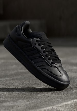 Czarny skórzany sneaker z teksturowaną podeszwą i czarnymi sznurowadłami, unoszący się w powietrzu ponad szarą betonową powierzchnią.