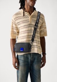 Black rectangular crossbody bag with zipper, blue logo patch, and adjustable strap. Model wears a beige knit polo and faded jeans.