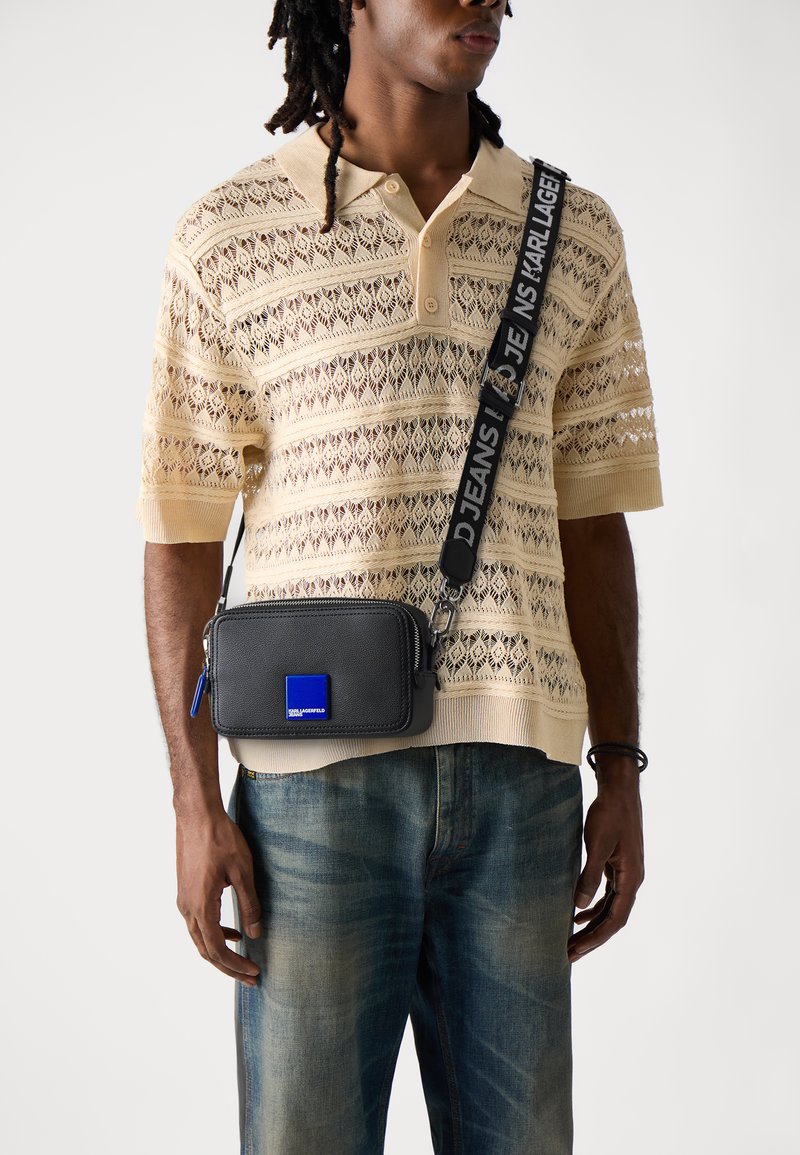 Black rectangular crossbody bag with zipper, blue logo patch, and adjustable strap. Model wears a beige knit polo and faded jeans.