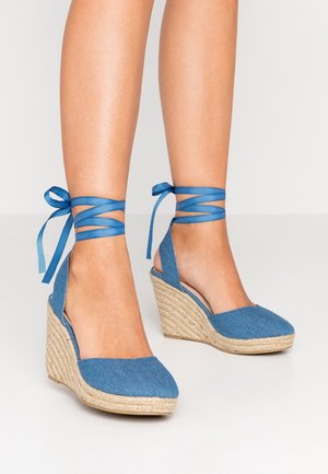 Benen met blauwe denim sleehak espadrilles met enkellintjes, gestrikt in strikjes, op een lichte achtergrond.