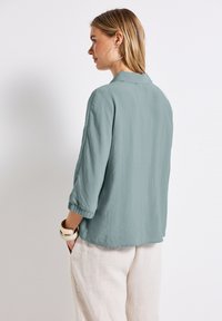 Camicetta azzurro chiaro, morbida e con maniche a tre quarti, colletto e tessuto testurizzato. Indossata con pantaloni di lino color crema.