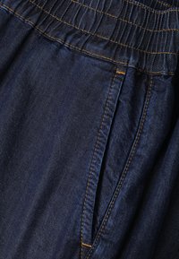 Κοντινή λήψη σκοτεινού μπλε denim υφάσματος που δείχνει ελαστική μέση και εμπρόσθια τσέπη με κίτρινο ράψιμο.