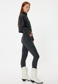 Czarna dżinsowa kurtka z srebrnymi guzikami, zestawiona z czarnymi obcisłymi dżinsami i białymi kowbojkami z dekoracyjnym przeszyciem. Model pozował z boku.