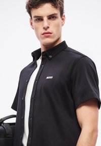 Μαύρο πουκάμισο με κοντά μανίκια και γιακά με κουμπιά, με διακριτικό λευκό λογότυπο "BOSS" στο στήθος, συνδυασμένο με λευκό εσώρουχο.