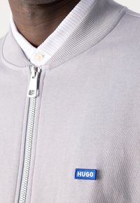 Nærbillede af person iført en lysegrå hættetrøje med ribbet krave over en hvid skjorte, med et lille blåt "HUGO" logo-patch.