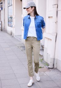 Femme marchant sur le trottoir, portant une chemise en jean bleue, un t-shirt blanc, un pantalon beige, des baskets blanches et une casquette à motif, souriante et regardant vers la gauche.