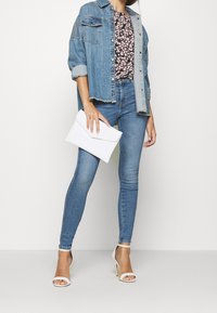 Denim jas over een bloemen top, gecombineerd met skinny jeans. Accessoires zijn een witte clutch en open-toe hakken in een lichte kleur.