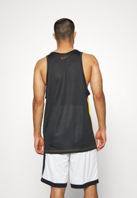 Débardeur de sport noir avec des panneaux latéraux jaunes, arborant un petit logo Nike orange à l'arrière. Porté avec un short blanc. Texture lisse.