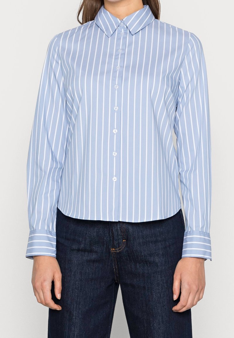 Lichtblauwe blouse met witte verticale strepen, lange mouwen en een puntige kraag. De stof lijkt glad en iets glanzend.