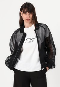 Black mesh bomber jacket with sheer panels and a ribbed collar, layered over a white t-shirt featuring black text, and paired with dark trousers.