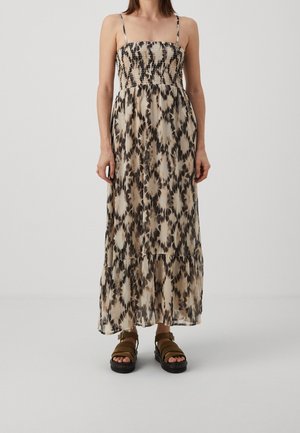 Vrouw in een beige en zwarte tie-dye maxi-jurk met dunne banden en bruine platform sandalen, staand tegen een effen grijze achtergrond.