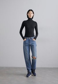 Top nero a collo alto, jeans chiari strappati con un buco al ginocchio e sneaker scure. La modella sta in piedi con le mani in tasca su uno sfondo neutro.